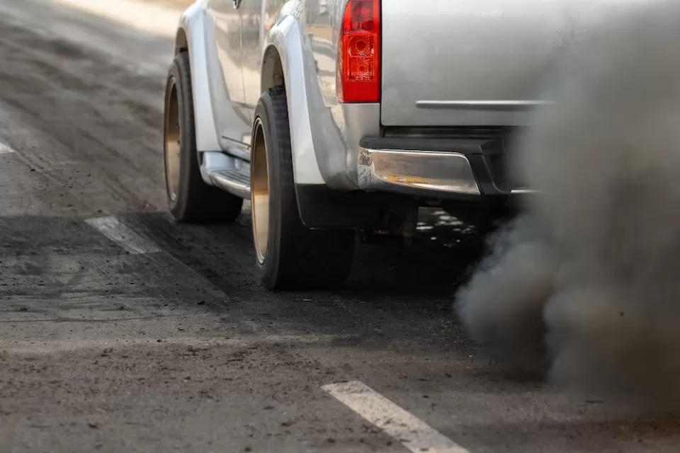 Close-up of driver's side of silver truck with cloud of dirty emissions from tailpipe.